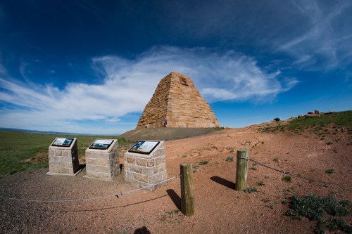 A stone pyramid structure on a grassy hill, with informational signs in the foreground and a blue sky above.