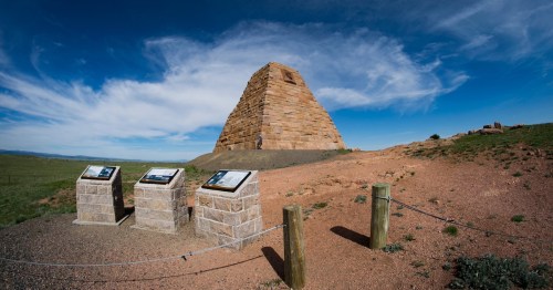 A stone pyramid structure on a grassy hill, with informational plaques in the foreground and a blue sky above.