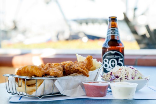 A basket of fried fish and fries, accompanied by coleslaw and dipping sauces, with a bottle of 805 beer in the background.