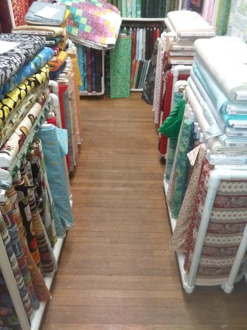 A narrow aisle between shelves filled with colorful fabric rolls in a craft store.