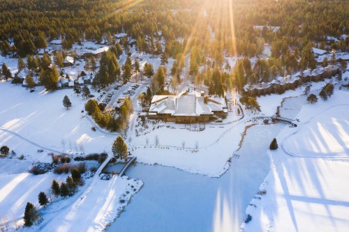 Aerial view of a snowy landscape with a lake, surrounded by trees and a cluster of buildings, illuminated by sunlight.
