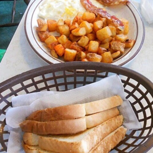 A plate with scrambled eggs, bacon, and seasoned potato cubes, alongside a basket of sliced bread.