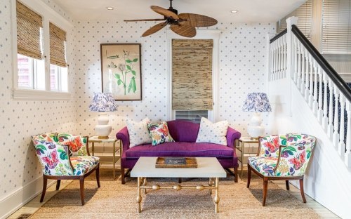 Bright living room with a purple sofa, floral chairs, a coffee table, and a ceiling fan, featuring light decor and natural light.