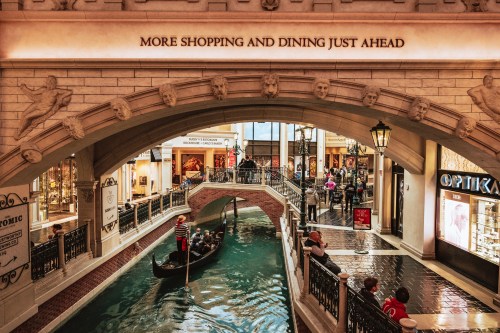 A scenic indoor canal with a gondola, surrounded by shops and dining areas, under an ornate archway.