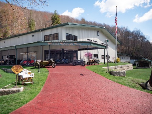 A building with a large glass front displays vintage motorcycles and vehicles, set against a green landscape and blue sky.