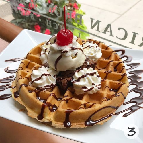 A delicious waffle topped with whipped cream, a cherry, chocolate drizzle, and a brownie piece on a white plate.