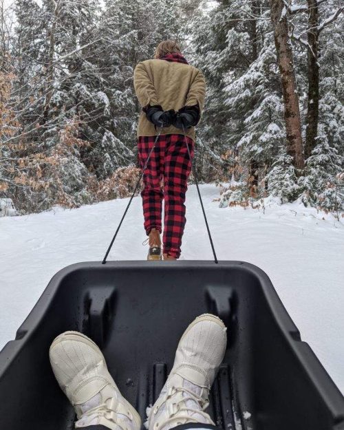 A person in a plaid outfit pulls a sled through a snowy forest, with winter boots visible in the foreground.