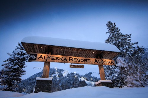 Sign for Alyeska Resort, Alaska, covered in snow, with a winter landscape and ski slopes in the background.