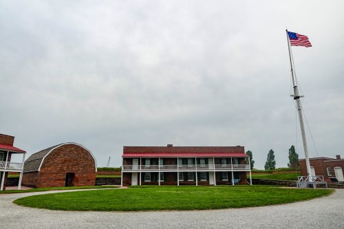 Historic buildings with a flagpole and American flag, set against a cloudy sky and green grass.