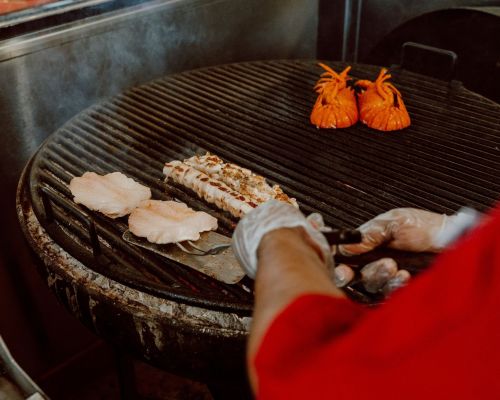 A chef grilling seafood, including fish fillets and lobster tails, on a large, smoky grill.