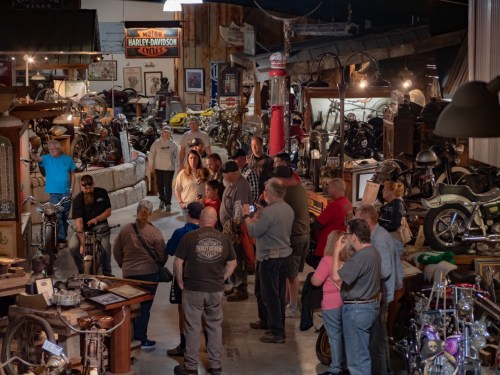 A crowd gathers in a vintage motorcycle museum, surrounded by various bikes and memorabilia.
