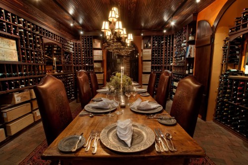 A cozy dining room with a large wooden table set for dinner, surrounded by wine racks filled with bottles.