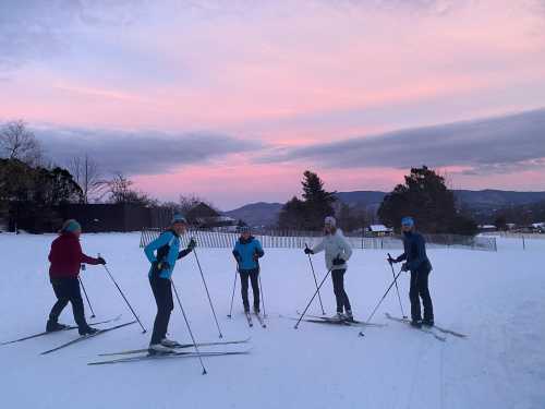 A group of five people cross-country skiing on a snowy landscape at sunset, with mountains in the background.