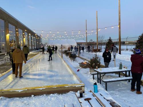 People enjoying an outdoor ice rink surrounded by snow, string lights, and festive decorations.