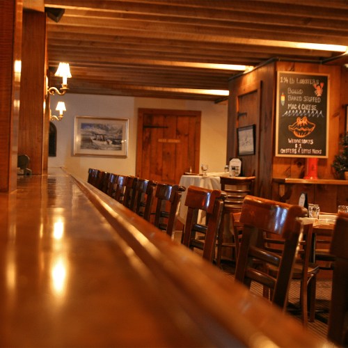 A rustic restaurant interior featuring wooden beams, a long bar, and empty tables with a chalkboard menu in the background.
