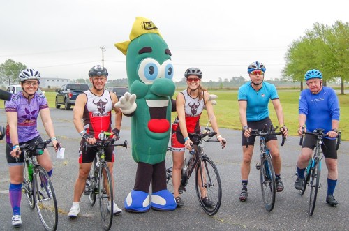 A group of five cyclists poses with a large pickle mascot in a rural setting, all smiling and ready to ride.