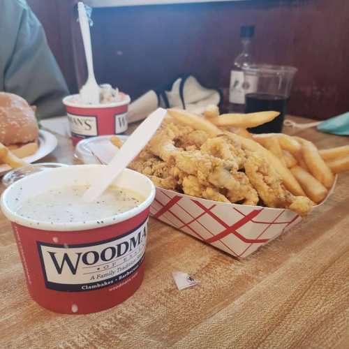 A bowl of clam chowder next to a tray of fried clams and French fries on a wooden table.