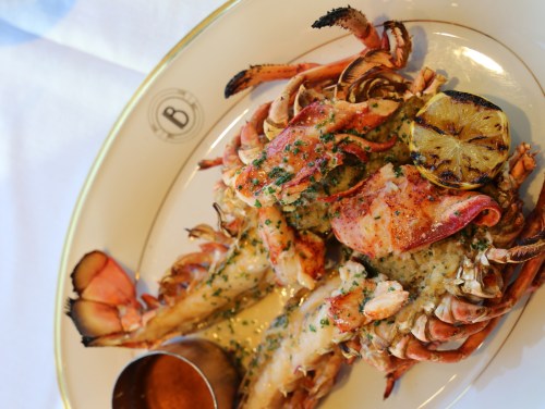 A beautifully plated lobster with grilled lemon and a small cup of sauce, garnished with herbs on a white tablecloth.