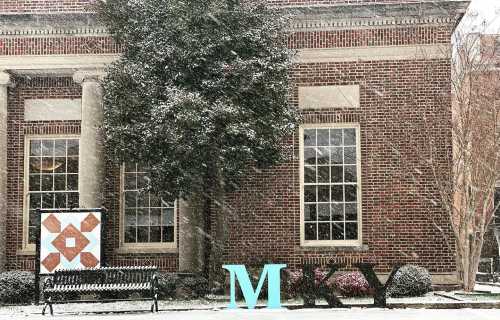 A snowy scene featuring a brick building, a bench, and colorful letters spelling "MKY" with a quilt pattern nearby.