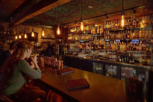 A cozy bar scene with a well-stocked shelf of bottles, warm lighting, and patrons enjoying drinks at the counter.