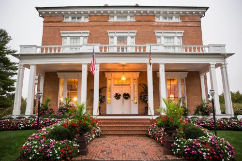 A large brick house with white columns, surrounded by colorful flower beds and American flags, on a cloudy day.