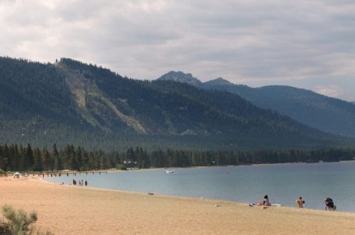 A serene beach scene with mountains in the background, people enjoying the sand and water under a cloudy sky.