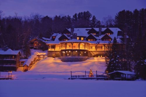 A beautifully lit lodge surrounded by snow, with a decorated Christmas tree in front and trees in the background at dusk.