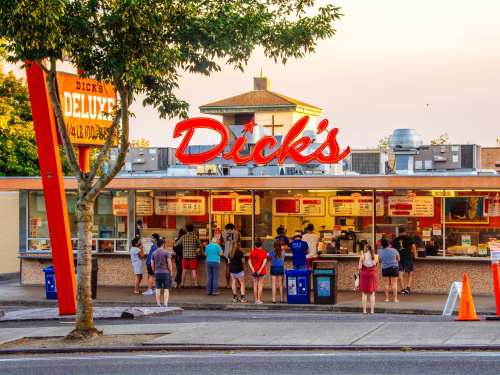 A busy fast-food restaurant with a bright sign, people lined up at the counter, and a warm sunset in the background.