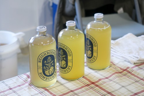 Three glass bottles of kombucha with labels reading "Neighborhood Kombuchery" on a checkered tablecloth.