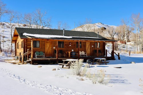 A rustic log cabin surrounded by snow, with a porch and chairs, set against a clear blue sky and distant hills.