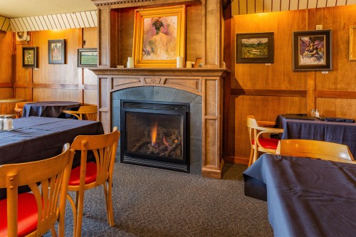 Cozy restaurant interior with wooden walls, a fireplace, and tables set with dark blue tablecloths. Artwork adorns the walls.