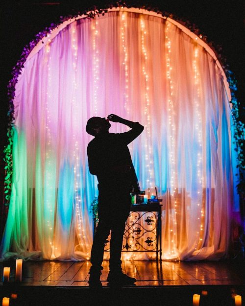 A silhouetted figure stands in front of a colorful, illuminated backdrop with draped fabric and fairy lights.