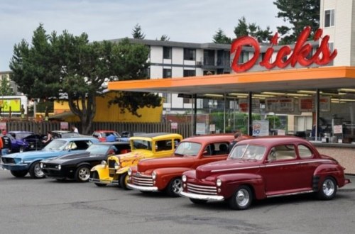 A row of classic cars parked in front of a diner with a bright orange sign reading "Dick's." Trees in the background.