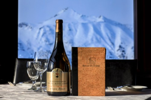 A bottle of wine and a menu sit on a table, with snow-capped mountains visible through a large window behind them.