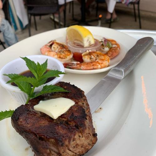 A plate featuring a grilled steak with butter and mint, alongside shrimp and lemon on a separate dish.