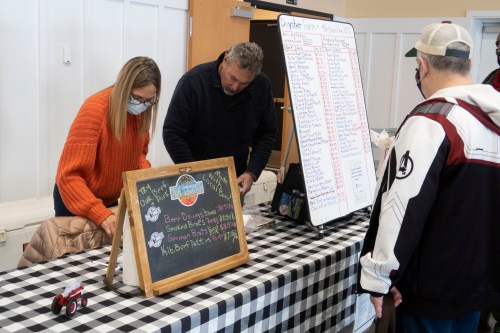 A woman and a man prepare food at a market stall, while a customer looks at the menu on a chalkboard.