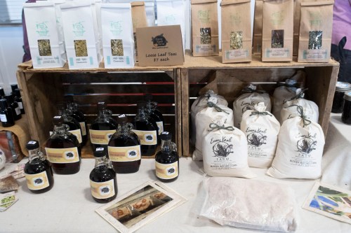 Display of herbal teas and natural products in bags and bottles on a wooden shelf at a market.