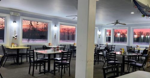 Cozy restaurant interior with empty tables, large windows showcasing a colorful sunset over a lake.
