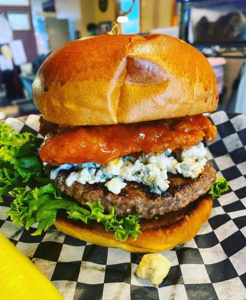 A stacked burger with a beef patty, crispy chicken, blue cheese, lettuce, and a toasted bun on a checkered paper.