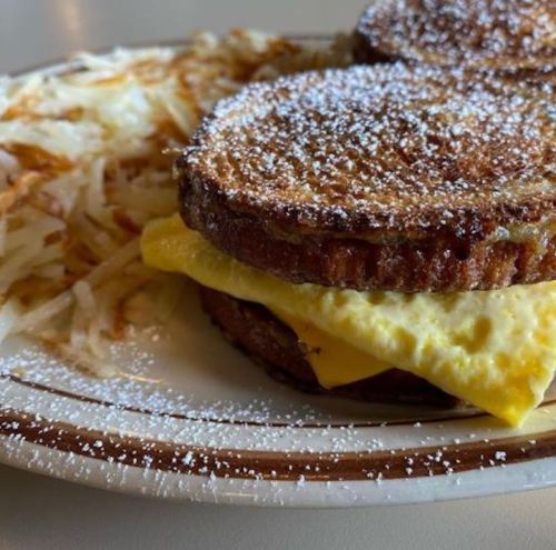 A plate with French toast sandwich filled with cheese and egg, served with crispy hash browns and powdered sugar on top.