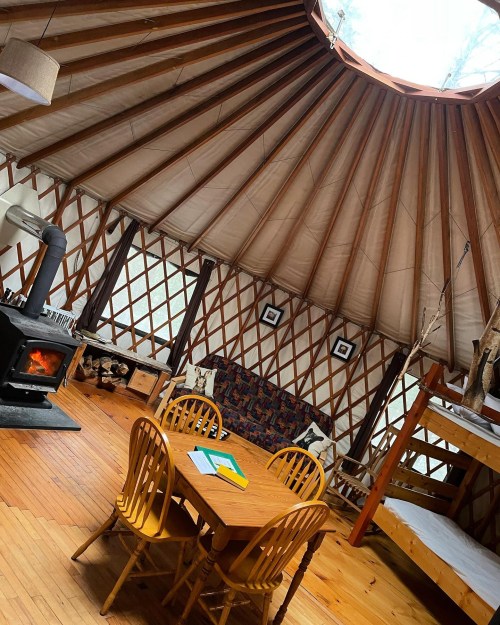 Interior of a yurt featuring a wooden table, chairs, a cozy seating area, and a wood stove. Natural light filters in.