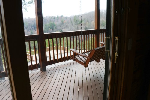 A wooden swing hangs on a porch overlooking a forested landscape, with a view of trees in the distance.