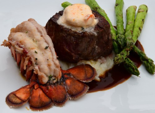 A gourmet dish featuring filet mignon, lobster tail, asparagus, and mashed potatoes drizzled with sauce.