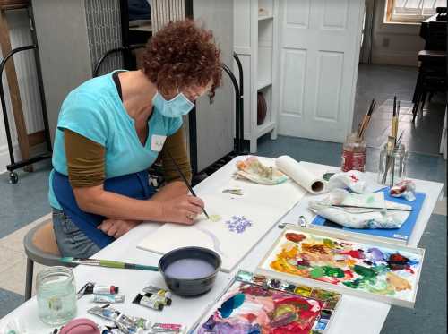 A woman in a mask paints on paper at a table filled with art supplies and colorful paint palettes.