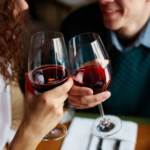 Two people clink wine glasses filled with red wine, smiling at each other in a cozy dining setting.