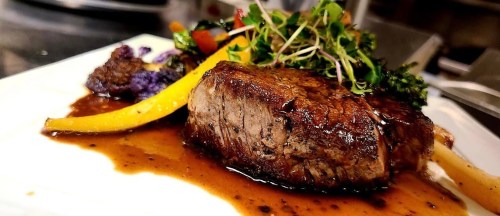 A beautifully plated steak with a rich sauce, garnished with microgreens and accompanied by colorful vegetables.