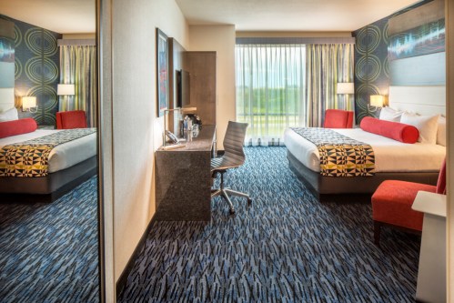 Modern hotel room featuring two beds, a desk, and large windows with natural light. Stylish decor and carpet.