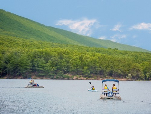 People in yellow life jackets paddle on a calm lake surrounded by green hills under a cloudy sky.
