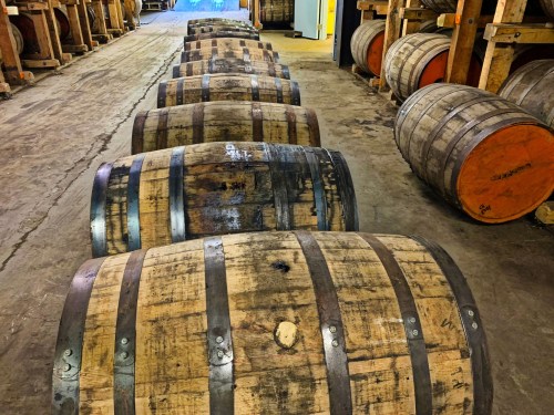 A row of wooden barrels lined up in a warehouse, with a rustic floor and soft lighting in the background.