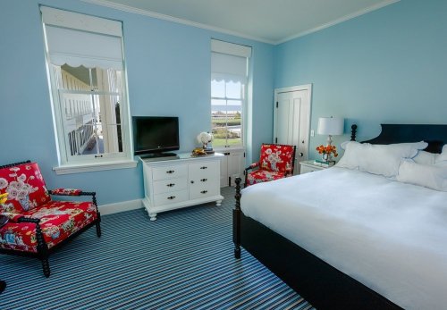 Bright bedroom with blue walls, a white bed, floral chairs, a TV, and large windows letting in natural light.
